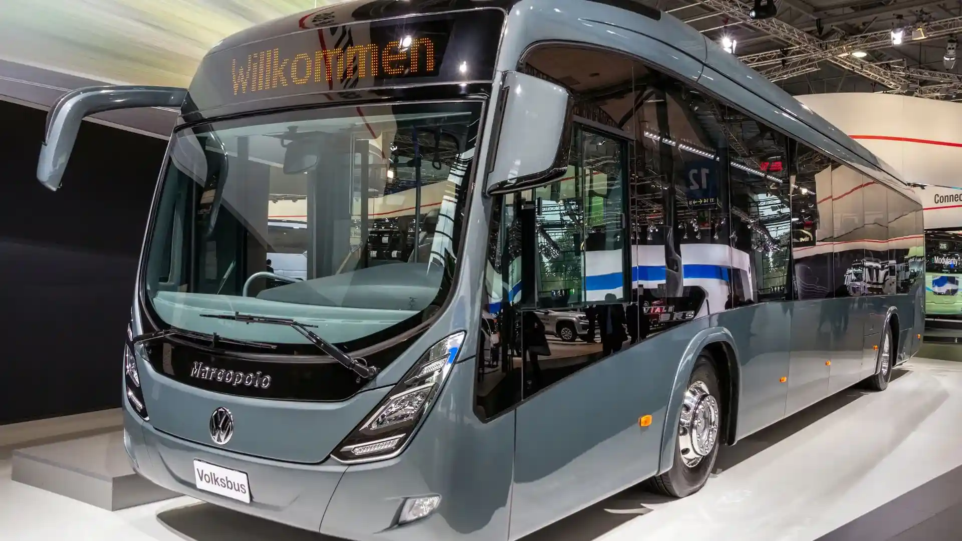 Ônibus Marcopolo Volksbus cinza em exposição, com fachada de vidro e letreiro ‘Willkommen’, apresentando design moderno de carroceria.