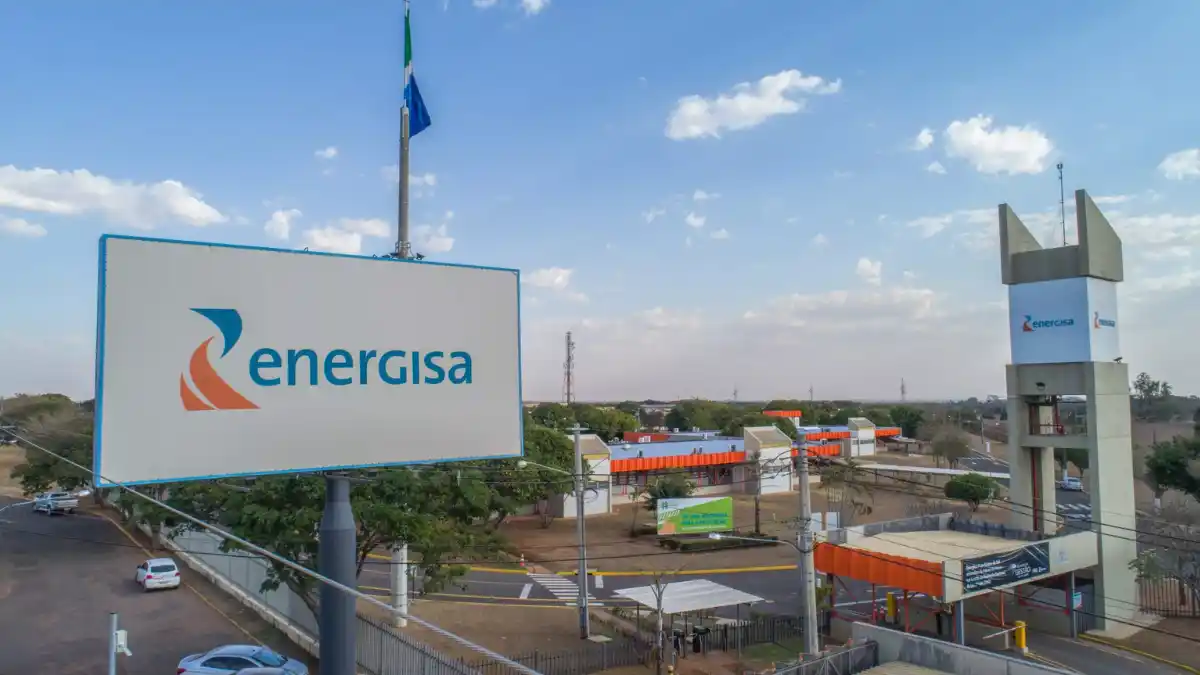Placa com o logotipo e o nome Energisa em frente a uma subestação ou planta de energia da empresa no setor elétrico.