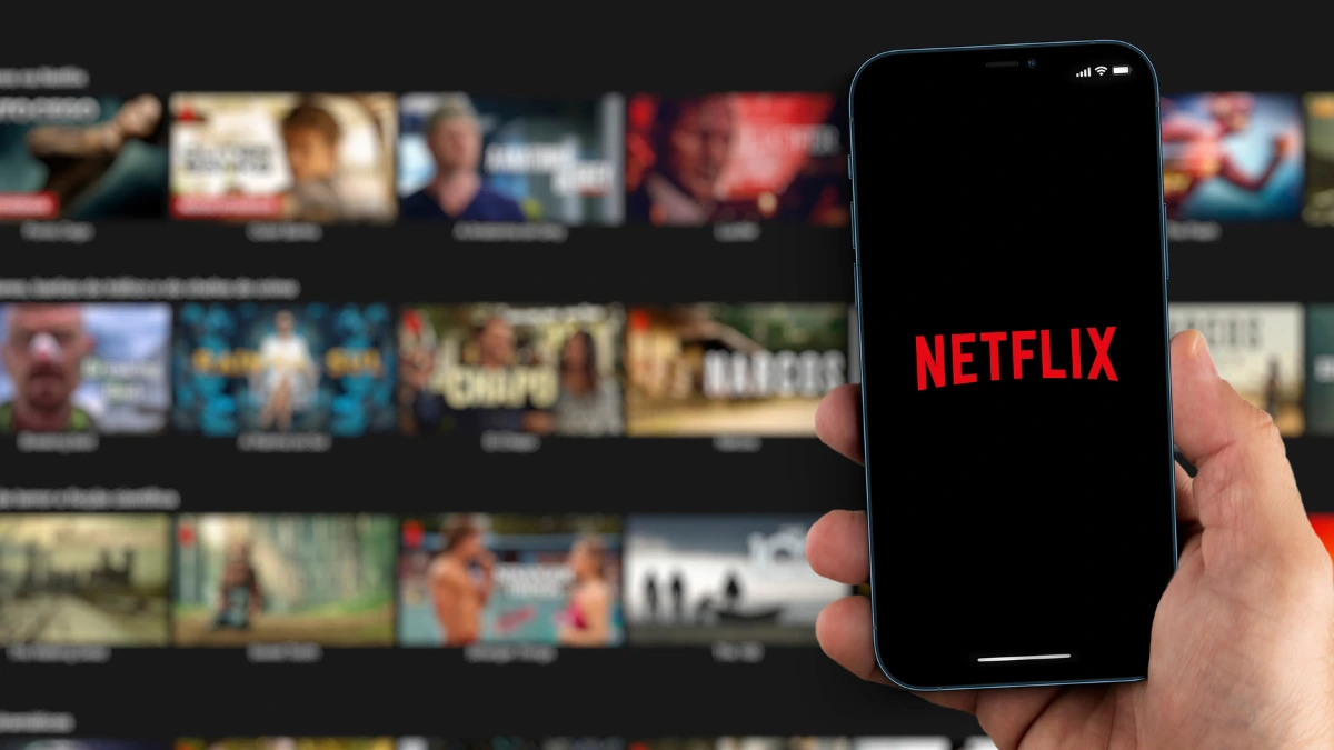 Mão segurando um smartphone que exibe o logotipo da Netflix (em primeiro plano), com a tela de conteúdo do aplicativo desfocada ao fundo.