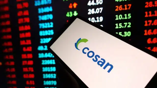 Smartphone exibindo o logotipo da Cosan em frente a um painel digital com cotações da bolsa de valores, representando a análise de mercado sobre a holding em 2026.