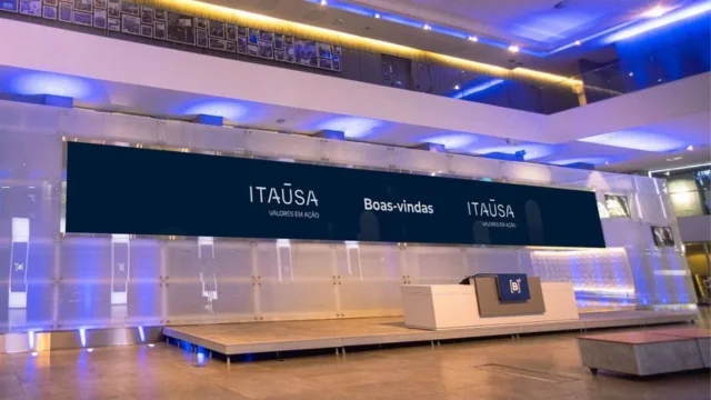 Grande grupo de executivos e colaboradores da Itaúsa comemorando no saguão da B3 sob chuva de confetes azuis e brancos, com o logotipo "Itaúsa 50 anos" no telão, ilustrando a solidez da itaúsa (itsa4) desconto de holding.
