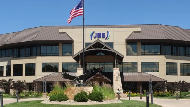 Fachada da sede corporativa da JBS nos Estados Unidos, um edifício de três andares com o logotipo da empresa e a bandeira americana hasteada à frente, ilustrando o contexto da jbs greve eua.