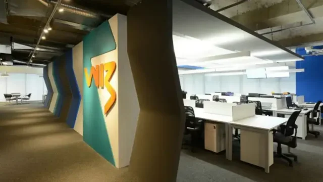 Placa metálica de parede com o logotipo da Wiz Co em relevo prateado sobre um fundo cinza texturizado, representando a sede da companhia para os resultados da Wiz 4T25.