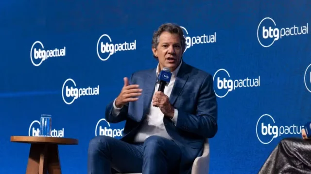 O Ministro Fernando Haddad discursando sentado em um painel durante evento corporativo, com o fundo azul exibindo logotipos do BTG Pactual.