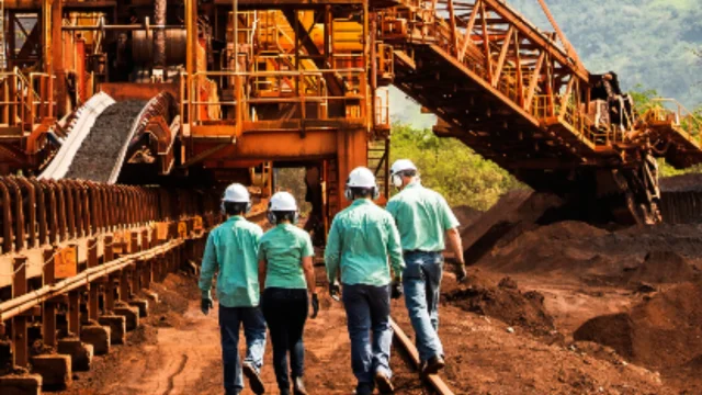 Quatro funcionários da Vale caminhando de costas em uma área de mineração com grandes estruturas de metal e correias transportadoras, representando o ativo Vale Thompson.