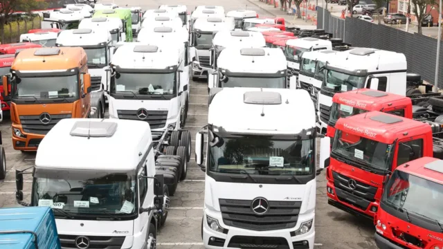 Vista aérea de um pátio lotado com dezenas de caminhões novos das marcas Mercedes-Benz e Atego, em cores branca, laranja e vermelha, enfileirados em um terminal logístico, ilustrando o setor afetado pela greve dos caminhoneiros.
