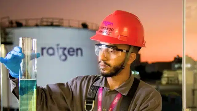 Profissional da Raízen com capacete vermelho e óculos de proteção analisando uma proveta com líquido azul em um laboratório industrial, com um tanque de armazenamento ao fundo, ilustrando o cenário de Raízen recuperação extrajudicial.