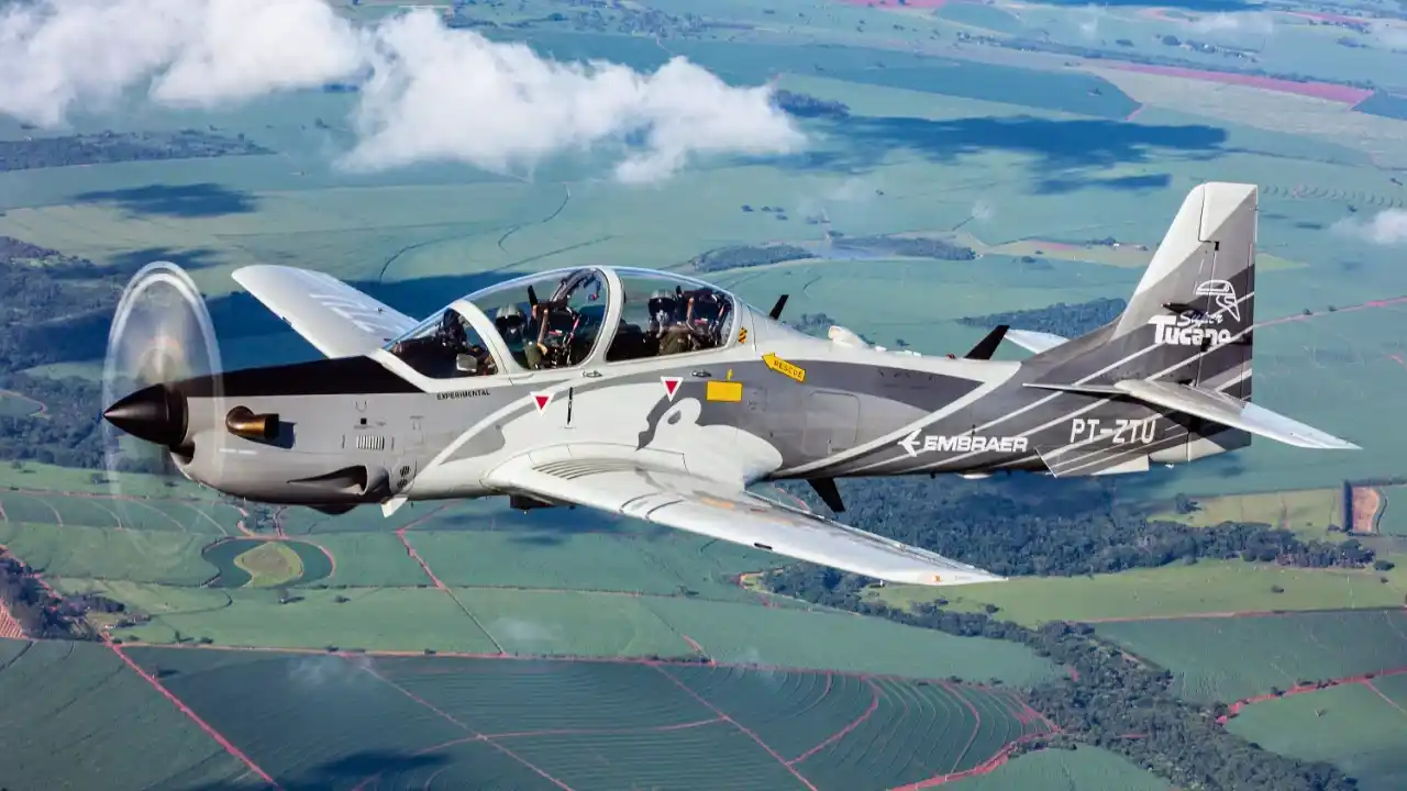 Vista lateral de uma aeronave Embraer Super Tucano em voo sobre uma paisagem rural verde com padrões de plantação. O avião possui pintura camuflada em tons de cinza e preto, hélice frontal em movimento e o logotipo da Embraer visível na cauda junto à identificação "Super Tucano" e o prefixo PT-ZTU.