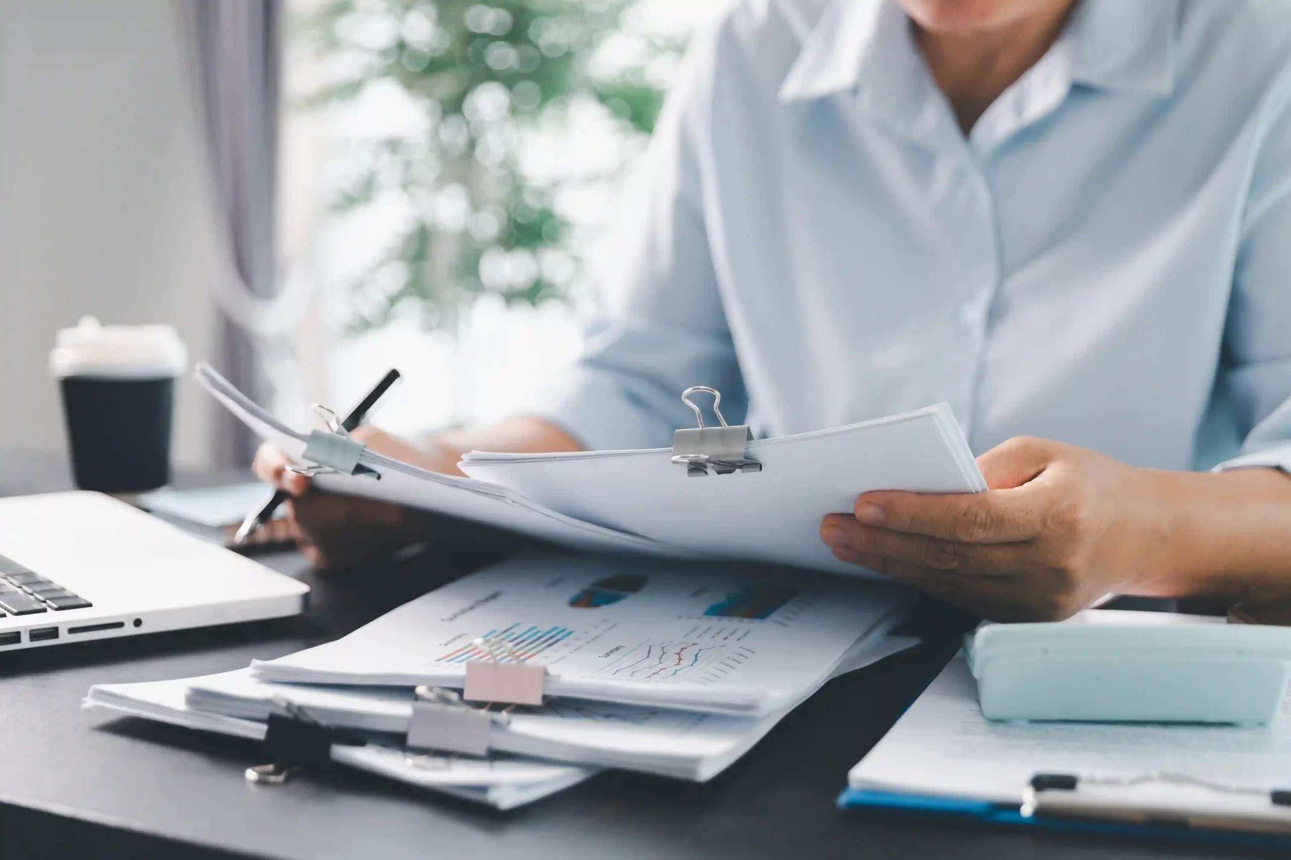 Mãos de um analista de finanças manuseando papéis e relatórios com gráficos e dados, simbolizando a gestão de portfólio de investimentos.