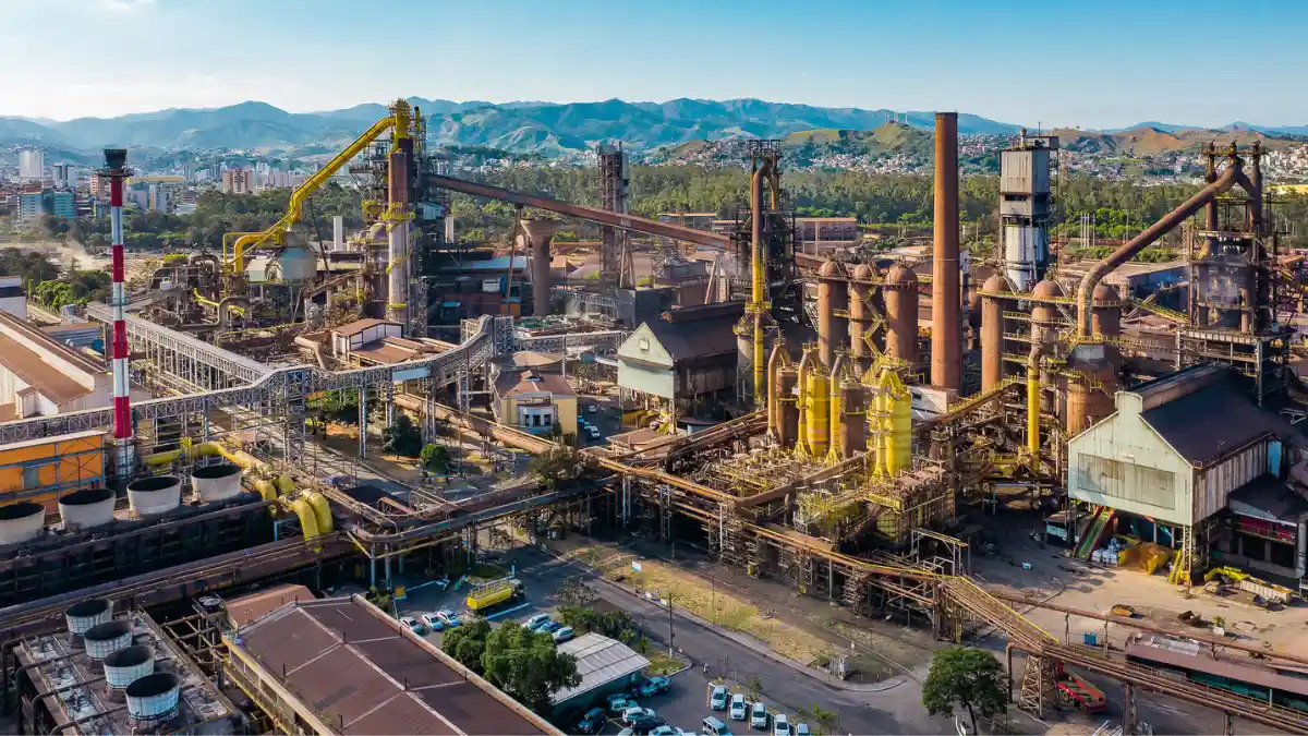 Vista aérea de uma grande usina siderúrgica da Usiminas, com fornos, chaminés e estruturas complexas da metalurgia, sob o horizonte da cidade.