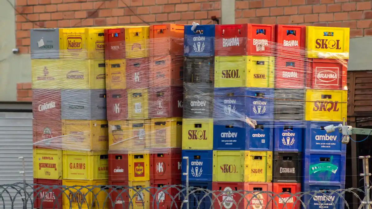 Pilha de caixotes de bebidas de diversas marcas da Ambev (Skol, Brahma, Budweiser, Stella Artois), simbolizando o balanço do 3º trimestre de 2025.