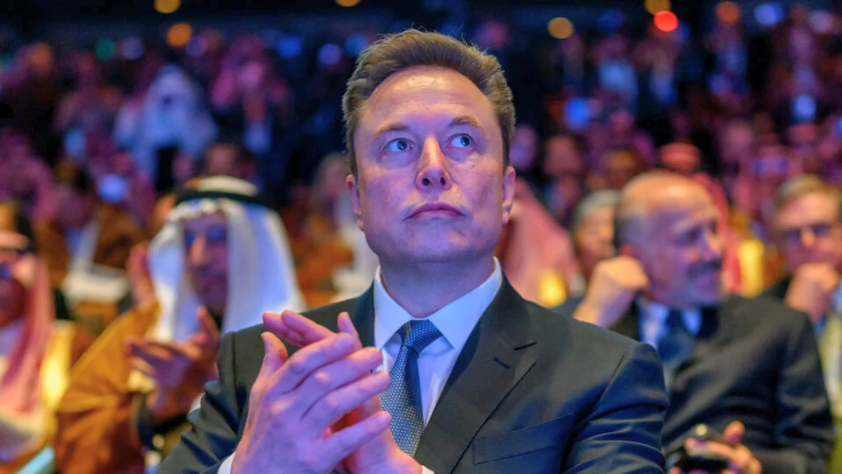 O empresário Elon Musk batendo palmas e olhando para cima durante um fórum, projetando que o trabalho pode se tornar opcional em 20 anos.