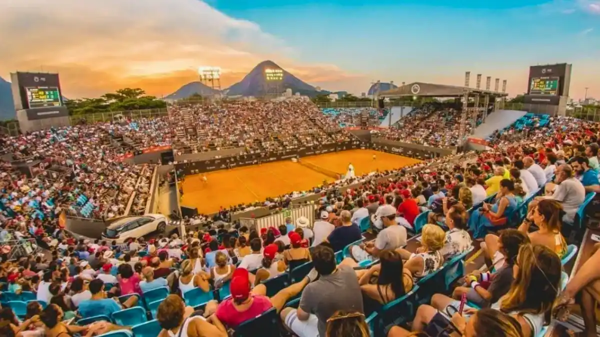 Vista aérea do complexo de tênis do Rio Open lotado de espectadores, com o Pão de Açúcar ao fundo, simbolizando o início da venda de ingressos para o evento.