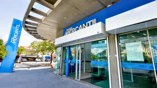 Fachada moderna de uma agência do Banco Mercantil com o logotipo azul em destaque acima da entrada de vidro, totem de identificação na calçada e arquitetura curva, representando o Banco Mercantil 2025.